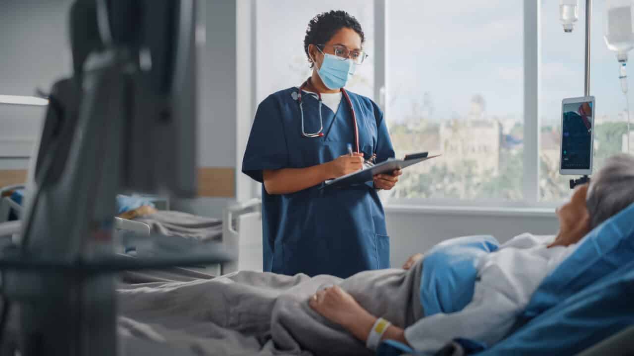 Hospital Ward: Friendly Nurse Wearing Face Mask Talks to Beautiful Senior Female Patient Resting in Bed, Checks Her Vitals, Writes down Information. Head Nurse and Old Lady Recovering after Covid 19